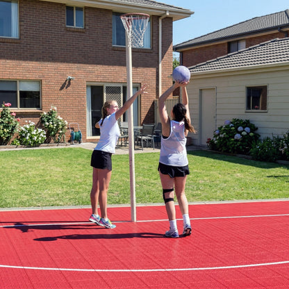 Two young women are playing basketball on a red court with a brick building and garden in the background.